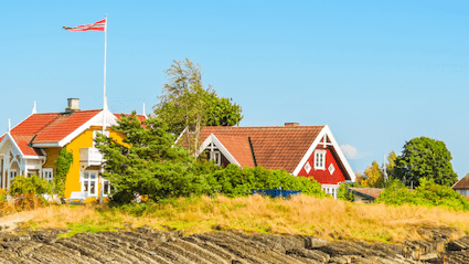 Et gult hus med hvite lister og norsk flagg på taket ved siden av et rødt hus med hvite lister omringet av trær og busker