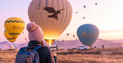 En kvinne med ryggen til kameraet, ser mot en klynge av varmluftsballonger i solnedgangen